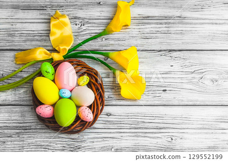 Colorful Easter eggs in a nest and yellow Calla flowers on a light wooden background. 129552199