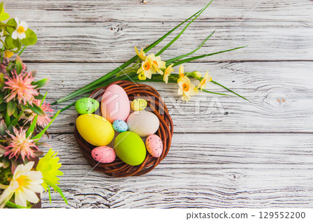 Colorful Easter eggs in the nest and narcissus flowers on light wooden background 129552200
