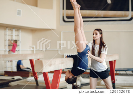Child gymnastics balance beam. Girl gymnast athlete during an exercise horizontal bar in gymnastics competitions. Coach with child. Child gymnastics balance beam. Girl gymnast athlete during an exercise horizontal bar in gymnastics competitions. Coach with child. 129552678