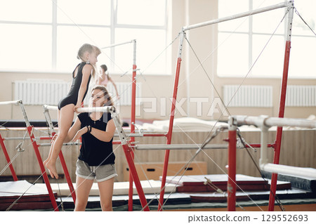 Child gymnastics balance beam. Girl gymnast athlete during an exercise horizontal bar in gymnastics competitions. Coach with child. Child gymnastics balance beam. Girl gymnast athlete during an exercise horizontal bar in gymnastics competitions. Coach with child. 129552693
