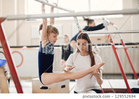 Child gymnastics balance beam. Girl gymnast athlete during an exercise horizontal bar in gymnastics competitions. Coach with child. 129552694