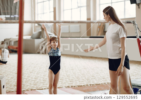 Child gymnastics balance beam. Girl gymnast athlete during an exercise horizontal bar in gymnastics competitions. Coach with child. Child gymnastics balance beam. Girl gymnast athlete during an exercise horizontal bar in gymnastics competitions. Coach with child. 129552695
