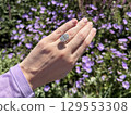 Landscape photo of woman's hand wearing a large oval cut diamond engagement ring, showcased against a blurred background of vibrant purple flowers in a sunlit garden. 129553308