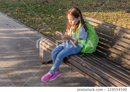Young girl with backpack using smartphone on park bench on a sunny day Young girl with backpack using smartphone on park bench on a sunny day 129554590
