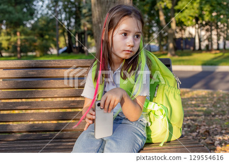 Child with smartphone and bright backpack sitting on park bench in autumn Child with smartphone and bright backpack sitting on park bench in autumn 129554616