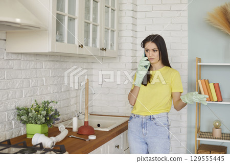Housewife woman standing in kitchen talking on phone to plumber service, wearing gloves Housewife woman standing in kitchen talking on phone to plumber service, wearing gloves 129555445