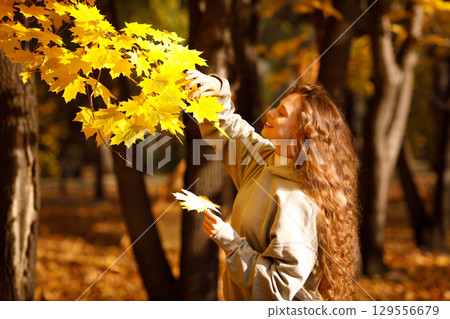 Smiling young woman enjoys the autumn weather in the forest with the yellow leaves at sunset 129556679