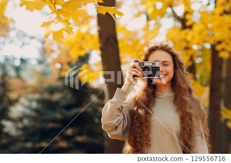 Smiling young woman with the photo camera shooting the forest trees with the yellow leaves at sunset Smiling young woman with the photo camera shooting the forest trees with the yellow leaves at sunset 129556716