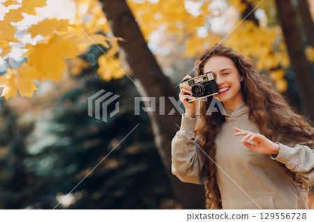 Smiling young woman photographer with the photo camera shooting the forest trees with the yellow leaves at sunset Smiling young woman photographer with the photo camera shooting the forest trees with the yellow leaves at sunset 129556728