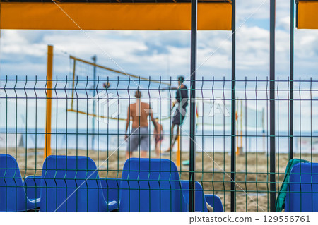 Beach Volleyball Court. View through fence of beach volleyball court with players and yellow net on sandy shore under cloudy sky. Concept of beach volleyball, summer sport, coastal outdoor activity Beach Volleyball Court. View through fence of beach volleyball court with players and yellow net on sandy shore under cloudy sky. Concept of beach volleyball, summer sport, coastal outdoor activity 129556761