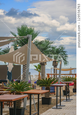 Outdoor Beach Cafe with Palm Trees, wooden tables, and decorative lights on sandy shore with cloudy sky. Concept of beach cafe, tropical seaside leisure, summer relaxation Outdoor Beach Cafe with Palm Trees, wooden tables, and decorative lights on sandy shore with cloudy sky. Concept of beach cafe, tropical seaside leisure, summer relaxation 129556763