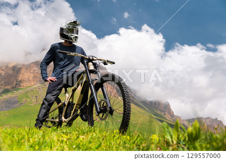 Cyclist standing with bicycle in fresh air. Tourist relaxing in field with hills view enjoying beautiful mountain landscape wearing helmet covering whole face. Active recreation concept Cyclist standing with bicycle in fresh air. Tourist relaxing in field with hills view enjoying beautiful mountain landscape wearing helmet covering whole face. Active recreation concept 129557000