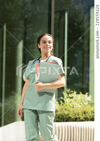 Smiling female doctor walking in the clinic yard with files in her hands Smiling female doctor walking in the clinic yard with files in her hands 129559228