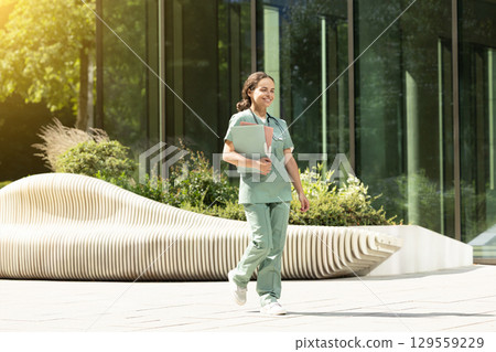 Smiling female doctor walking in the clinic yard with files in her hands 129559229