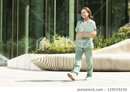 Smiling female doctor walking in the clinic yard with files in her hands 129559230