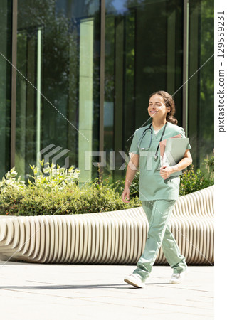 Smiling female doctor walking in the clinic yard with files in her hands 129559231