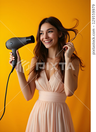 Joyful woman drying her hair with a hairdryer against a bright yellow backdrop in a stylish setting 129559979