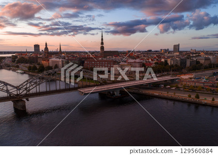 Riga's skyline at sunset with the Daugava River, metal railway bridge, modern road bridge, and St. Peter's Church spire in the historic old town. 129560884