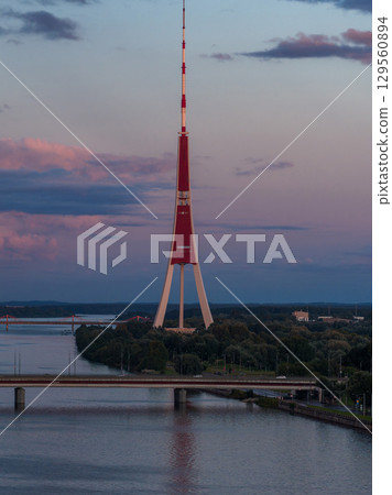 The Riga TV Tower stands tall against a sunset sky, framed by greenery and the Daugava River. A bridge spans the river, reflecting in the calm water. The Riga TV Tower stands tall against a sunset sky, framed by greenery and the Daugava River. A bridge spans the river, reflecting in the calm water. 129560894