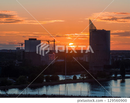 Warm sunset over Riga, Latvia, featuring the Swedbank building by the Daugava River, a Latvian flagpole, and construction cranes in the background. Warm sunset over Riga, Latvia, featuring the Swedbank building by the Daugava River, a Latvian flagpole, and construction cranes in the background. 129560900