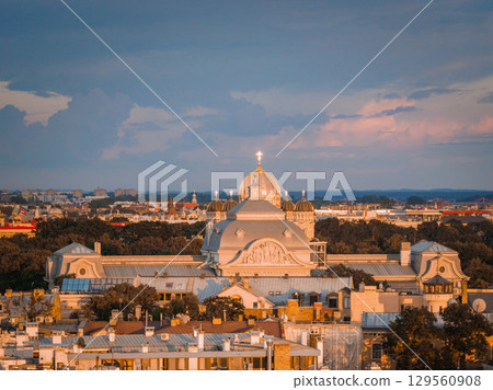 Golden sunset light illuminates the Nativity of Christ Cathedral in Riga, Latvia, with its domes and cross, surrounded by rooftops and a tree lined background. 129560908