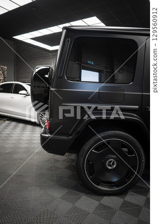 Black Mercedes AMG G Class SUV rear view with spare tire cover and alloy wheels. Indoor showroom setting with white luxury car in the background. Black Mercedes AMG G Class SUV rear view with spare tire cover and alloy wheels. Indoor showroom setting with white luxury car in the background. 129560912