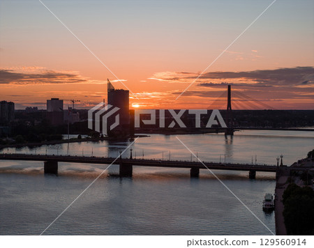 A sunset view of Riga, Latvia, featuring the Daugava River, a bridge with streetlights, the Riga TV Tower, and a modern high rise near the riverbank. 129560914