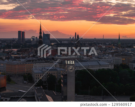 Riga's old town spires, including St. Peter's Church and Riga Cathedral, stand against a vibrant orange and pink sunset, with a clock tower in the foreground. 129560917