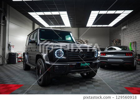A black Mercedes AMG G Class with illuminated LED headlights and a gray Ford Mustang GT 5.0 in a well lit garage with checkered flooring. A black Mercedes AMG G Class with illuminated LED headlights and a gray Ford Mustang GT 5.0 in a well lit garage with checkered flooring. 129560918