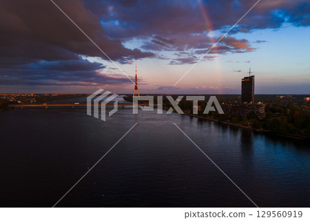 View of Riga, Latvia, featuring the Riga TV Tower, a faint rainbow, the Daugava River, a bridge, modern buildings, and dramatic sunset clouds. 129560919