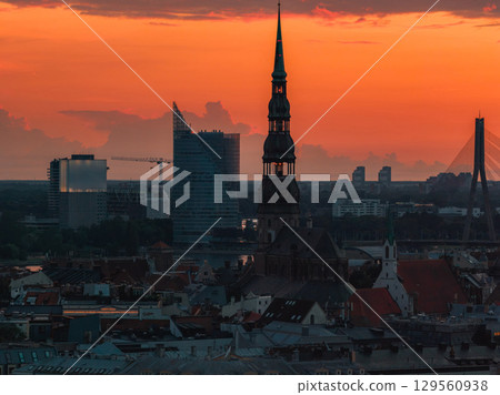 Silhouette of St. Peter's Church spire at sunset in Riga, Latvia, with old town rooftops, Vansu Bridge, and modern buildings in the background. 129560938