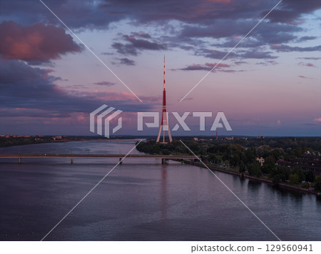 Riga TV Tower stands on an island in the Daugava River at sunset, with a bridge in the foreground, lush greenery, and soft pink and purple skies. Riga TV Tower stands on an island in the Daugava River at sunset, with a bridge in the foreground, lush greenery, and soft pink and purple skies. 129560941