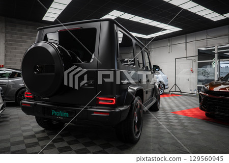 Matte black Mercedes AMG G Class SUV parked indoors on a checkered floor. Rear lights, spare tire cover, and sleek design visible in a bright showroom. 129560945