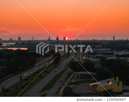 A sunset view of Riga, Latvia, featuring the old town spires, Riga TV Tower, Daugava River, a bridge, and a tree lined road with vehicles. A sunset view of Riga, Latvia, featuring the old town spires, Riga TV Tower, Daugava River, a bridge, and a tree lined road with vehicles. 129560947