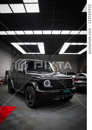 A sleek black Mercedes AMG G Class with illuminated LED headlights displayed in a showroom with a grid patterned ceiling and checkered flooring. A sleek black Mercedes AMG G Class with illuminated LED headlights displayed in a showroom with a grid patterned ceiling and checkered flooring. 129560950
