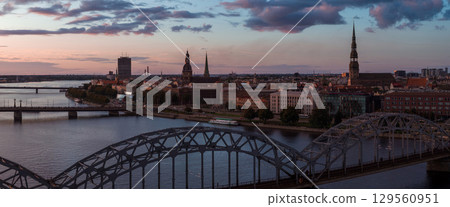 Riga, Latvia at sunset with the Daugava River, arched railway bridge, Riga Cathedral, and St. Peter's Church spire under a warm, cloud filled sky. 129560951