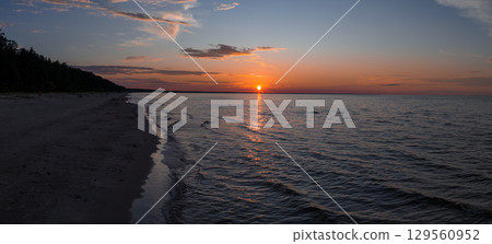 A tranquil sunset over calm waters in Jurmala, Latvia, with a sandy beach on the left and a dense line of trees under a warm orange sky. 129560952