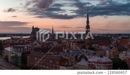 Riga's old town skyline at sunset features St. Peter's Church spire, Riga Cathedral, and Jugendstil architecture with the Daugava River in the background. 129560957