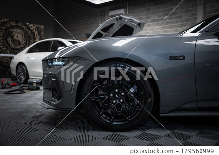 Gray Ford Mustang GT 5.0 with black alloy wheels and Brembo brakes in focus. White Mercedes AMG G Class with open hood in an industrial setting. Gray Ford Mustang GT 5.0 with black alloy wheels and Brembo brakes in focus. White Mercedes AMG G Class with open hood in an industrial setting. 129560999