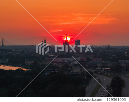 Sunset over Riga, Latvia, with the Latvian Academy of Sciences spire silhouetted, Vansu Bridge over the Daugava River, and lush greenery in the foreground. 129561003