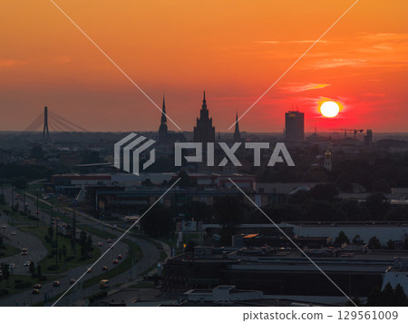 Silhouettes of Riga's old town spires, Vansu Bridge, and urban structures under a warm orange sunset sky, with modern and historical elements visible. Silhouettes of Riga's old town spires, Vansu Bridge, and urban structures under a warm orange sunset sky, with modern and historical elements visible. 129561009