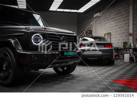 Black Mercedes AMG G Class with circular LED headlights and gray Ford Mustang GT 5.0 in a modern workshop with tiled floor and industrial walls. Black Mercedes AMG G Class with circular LED headlights and gray Ford Mustang GT 5.0 in a modern workshop with tiled floor and industrial walls. 129561014