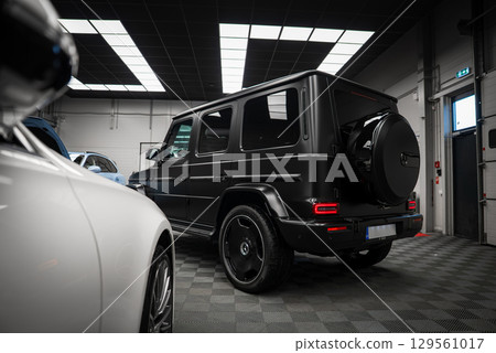 Sleek black Mercedes AMG G Class SUV with matte finish on checkered floor, rear and side profile visible, under bright grid patterned lights. 129561017