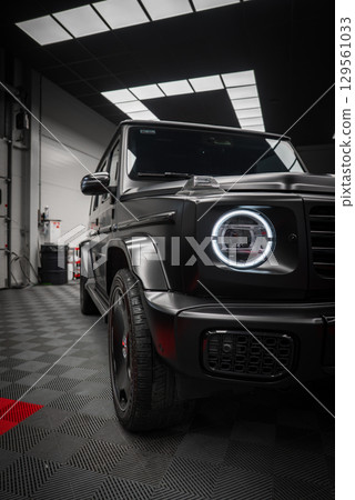 Black Mercedes AMG G Class SUV with matte finish, circular LED headlight, and bold grille in a modern showroom with patterned black floor. 129561033