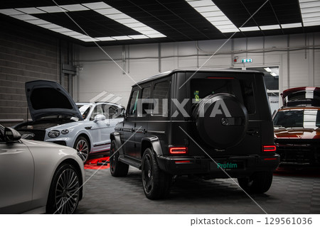 A black Mercedes AMG G Class SUV displayed indoors, showcasing its boxy rear design, spare tire cover, and taillights. Other luxury cars surround it. 129561036