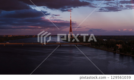 Riga TV Tower stands tall during sunset with purple and pink skies. The Daugava River flows below, crossed by a bridge with light vehicle traffic. Riga TV Tower stands tall during sunset with purple and pink skies. The Daugava River flows below, crossed by a bridge with light vehicle traffic. 129561039