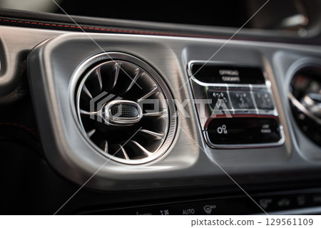 Close up view of a Mercedes AMG G Class interior, highlighting a turbine style air vent, red stitched dashboard trim, and sleek climate controls. 129561109