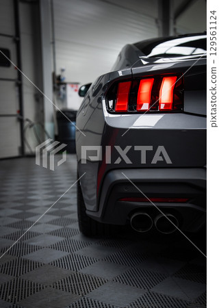 Rear view of a gray Ford Mustang GT 5.0 with illuminated triple taillights, dual exhaust, and sleek lines in a showroom with checkered flooring. Rear view of a gray Ford Mustang GT 5.0 with illuminated triple taillights, dual exhaust, and sleek lines in a showroom with checkered flooring. 129561124