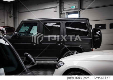 A sleek black Mercedes AMG G Class SUV with a matte finish parked indoors. Features include its boxy design, rear spare tire, and polished surroundings. 129561132