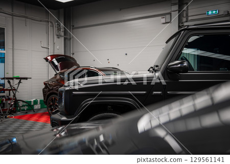 A black Mercedes AMG G Class in the foreground and a red Ford Mustang GT 5.0 with its hood open in a modern indoor showroom with tiled flooring. A black Mercedes AMG G Class in the foreground and a red Ford Mustang GT 5.0 with its hood open in a modern indoor showroom with tiled flooring. 129561141